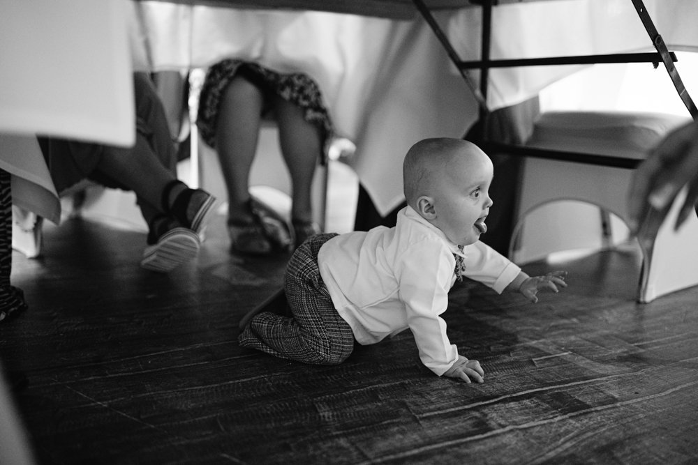 A baby in a white shirt and plaid pants crawls on a wooden floor under a table. People are seated at the table with legs and shoes visible.