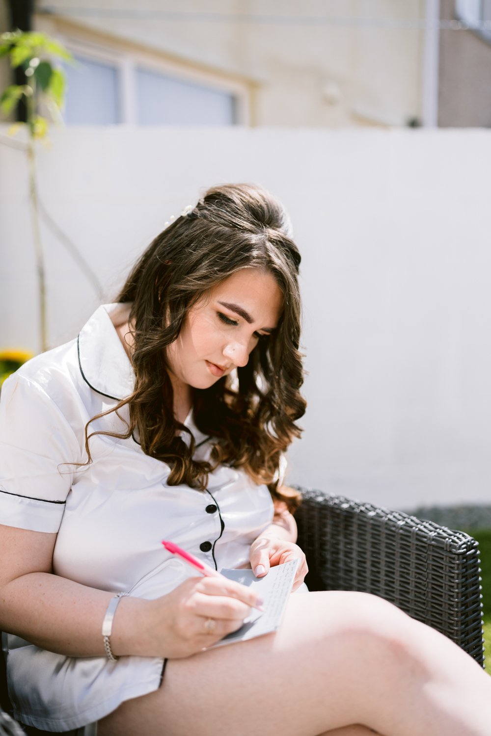 Woman sitting outdoors in a wicker chair, wearing a white shirt with black trim, writing in a notepad with a red pen, with a white wall and plants in the background.