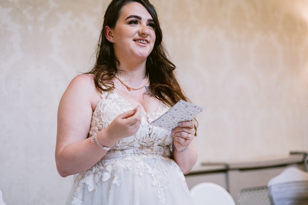 Woman in a lace dress gives a speech, holding note cards, against a light floral-patterned wall.