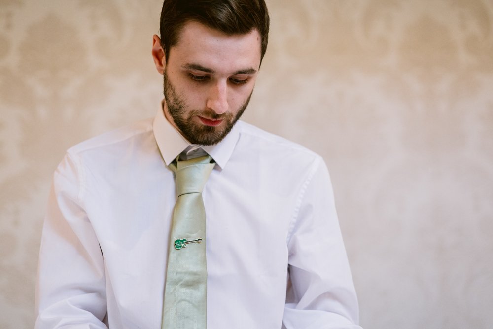 A man with a beard wearing a white shirt and light green tie with a guitar-shaped pin stands against a patterned beige background.