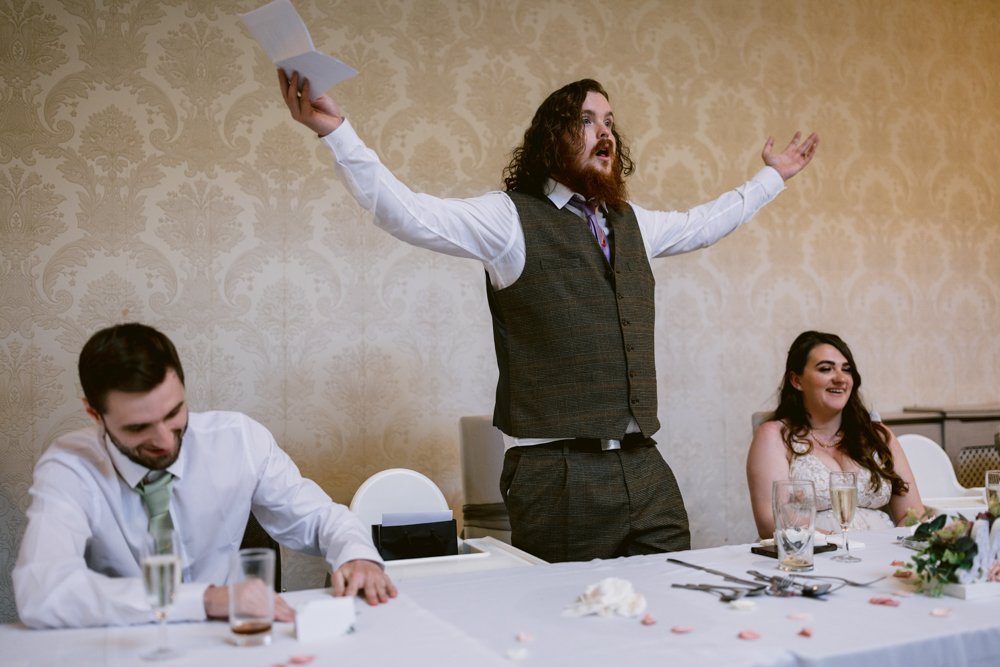 A man in a suit stands with arms outstretched, holding a piece of paper, while two people seated beside him, a man and a woman, smile at a table with drinks and decorations.