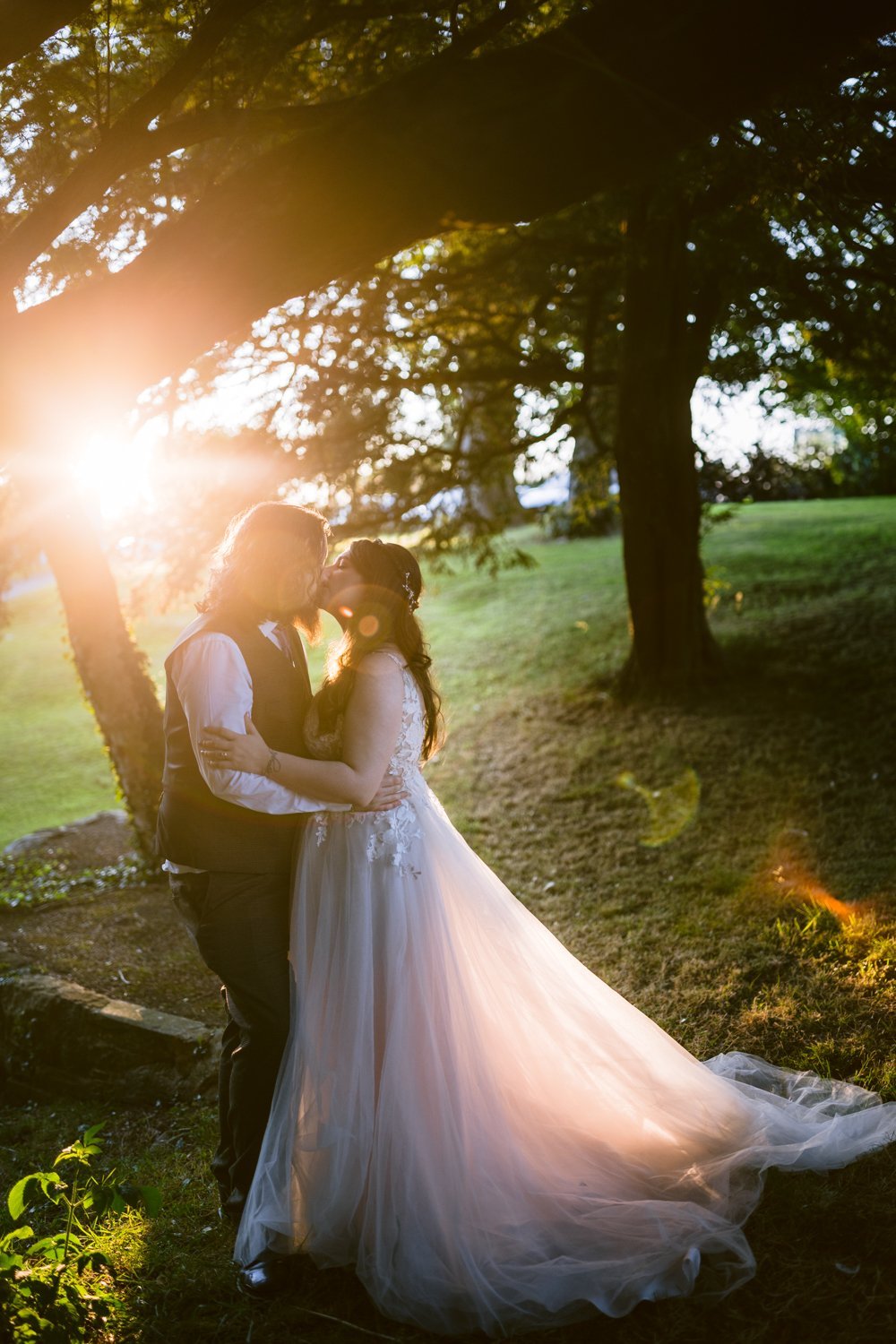 A couple in wedding attire embraces under trees with sunlight streaming through, casting warm, glowing highlights around them.
