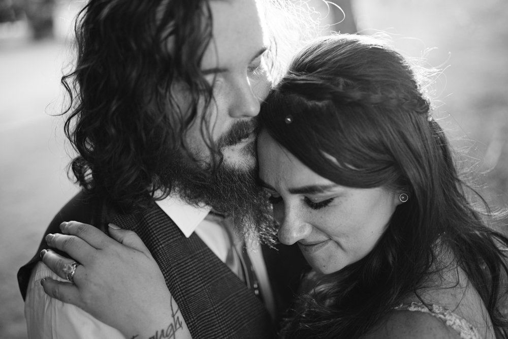 A black and white photo of a couple embracing closely. The man has long hair and a beard, wearing a vest. The woman has long hair and closes her eyes, resting her head on his chest.