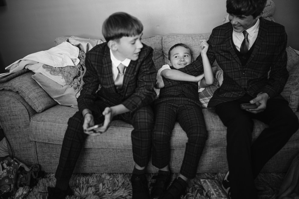 Three children in suits sit on a couch. The child in the middle is smiling and leaning back, while the others are holding devices. The scene is in black and white.