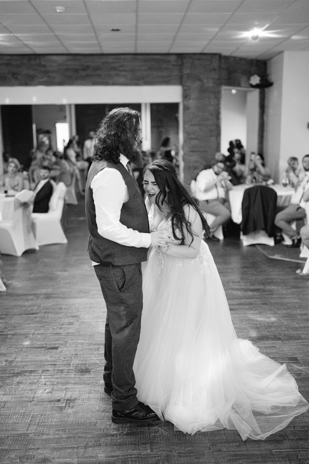 Bride and groom dancing in a large room, surrounded by seated guests. The bride is smiling widely. The scene is black and white.