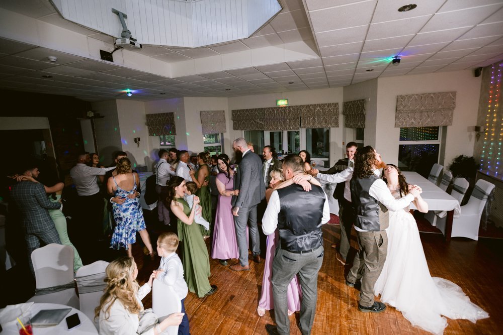A wedding reception dance floor with guests dancing and chatting. The room features white chairs and decorated tables. A bride and groom are dancing in the foreground.