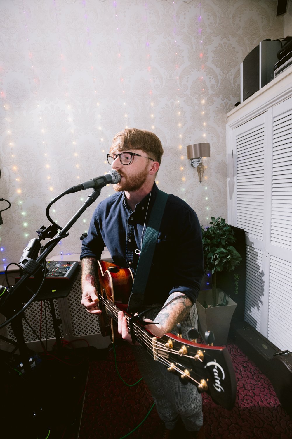 Person singing and playing an acoustic guitar in a room with decorative lights and wallpaper.