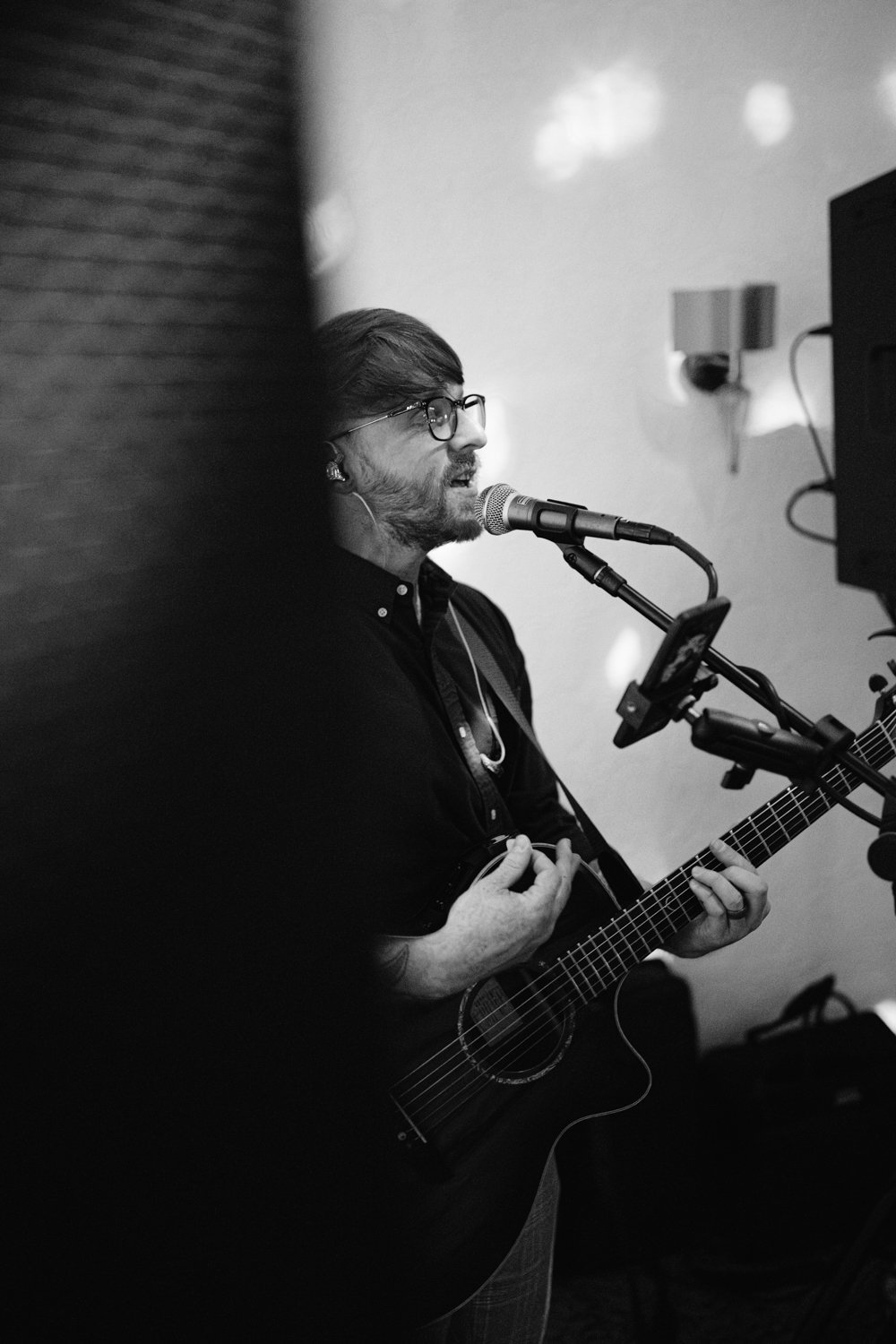 A person with glasses is playing an acoustic guitar and singing into a microphone. The scene is indoors, with studio lights and equipment visible.
