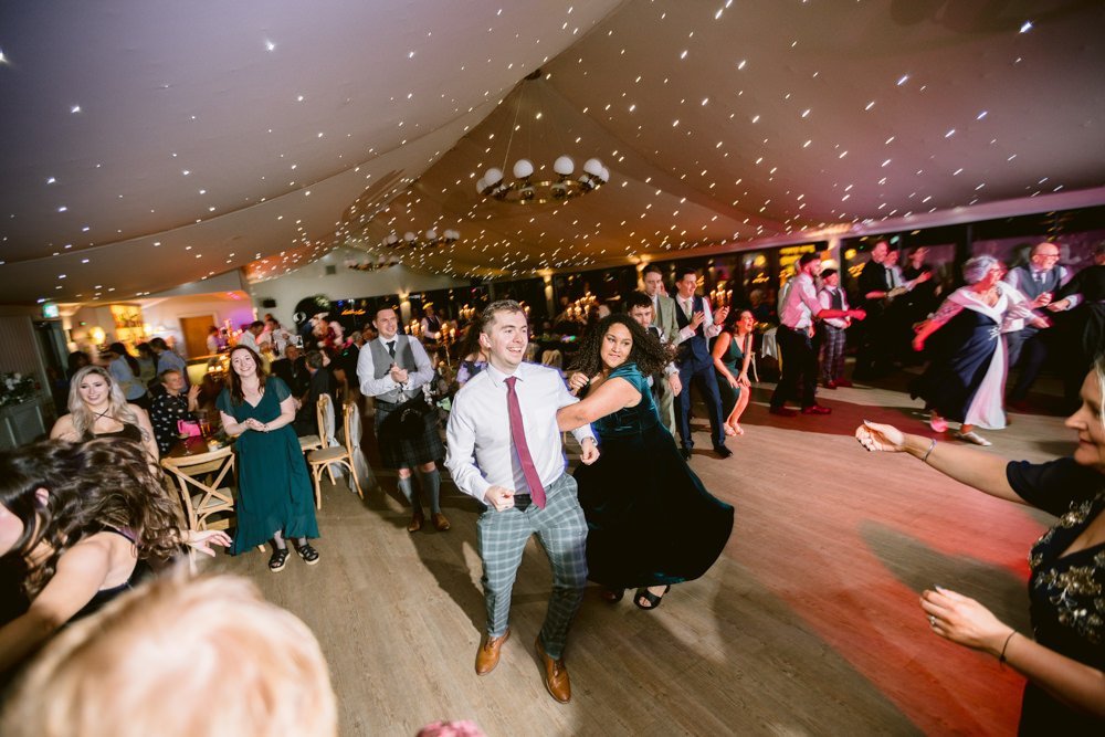 People dancing energetically at a party in a star-lit room with a wooden floor and tables in the background.