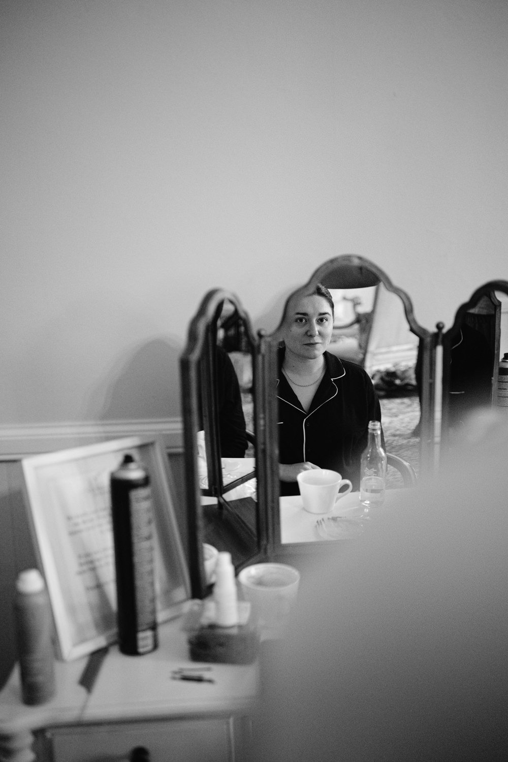 A person in pajamas is reflected in a tri-fold mirror on a dresser. The dresser holds various items, including a bottle, cup, and spray can. The scene is in black and white.