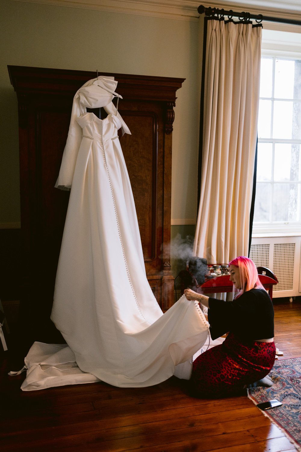 A person with pink hair steams a long white wedding dress hanging on an armoire in a room with wooden floors and large windows.