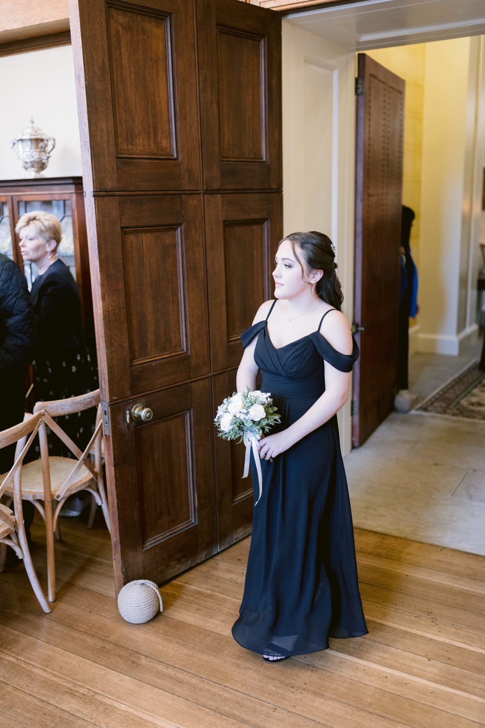 A woman in a black dress holds a small bouquet while standing in a doorway at an indoor event.