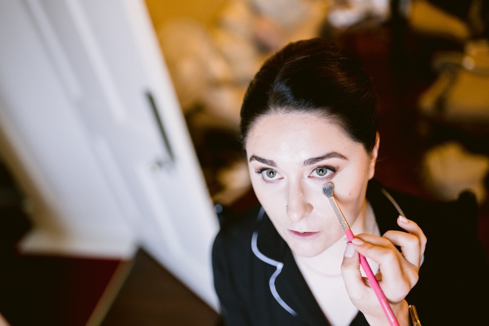 Person getting makeup applied with a brush, wearing a black top, in a softly lit room.