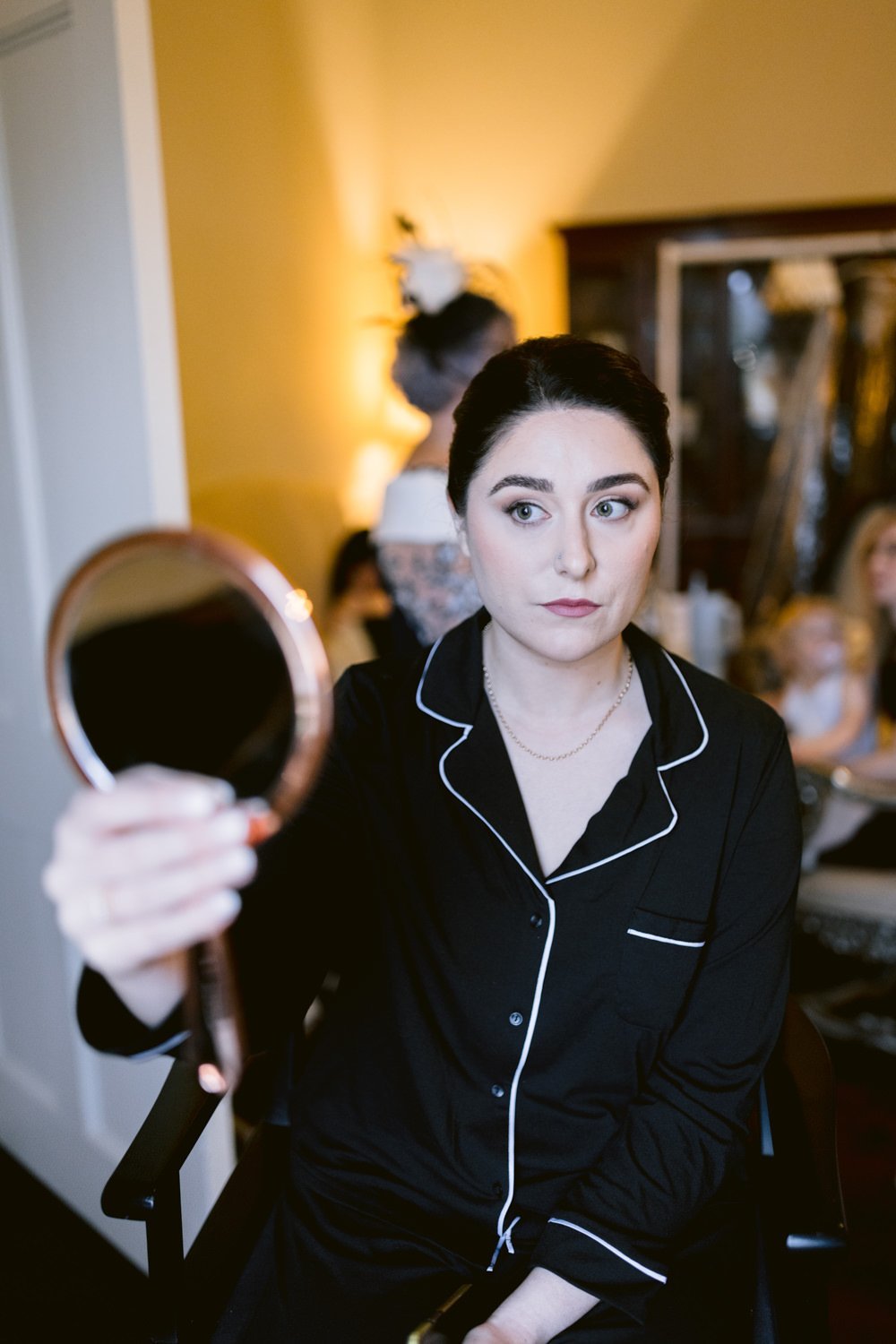 A person in black pajamas holds a handheld mirror, looking into it with a neutral expression. A blurred background shows figures and a warm-colored wall.