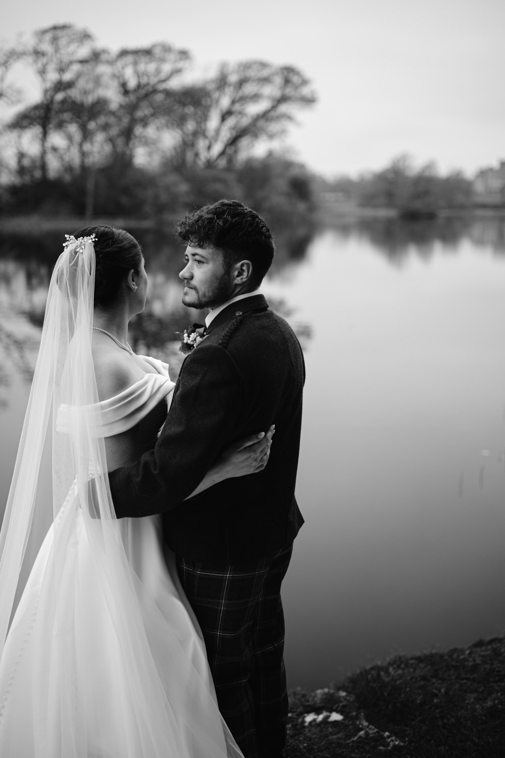 A bride and groom embrace near a calm lake, surrounded by trees. The photo is in black and white.