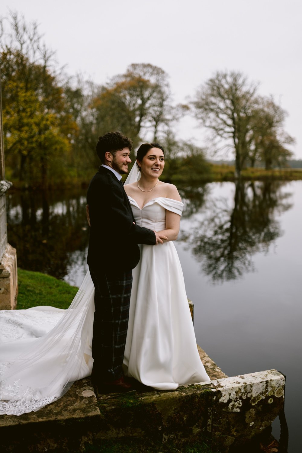 A couple in wedding attire stands on a stone platform by a reflective lake, surrounded by autumn trees.