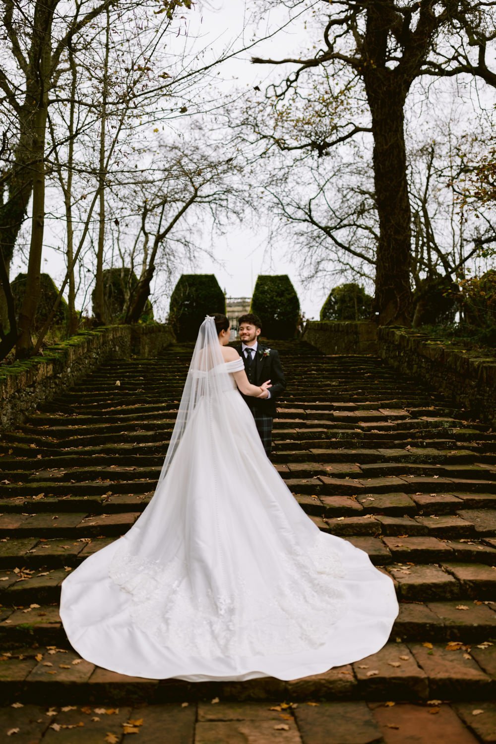 Bride and groom standing on stone steps in a wooded area, with the bride wearing a long white gown and veil.