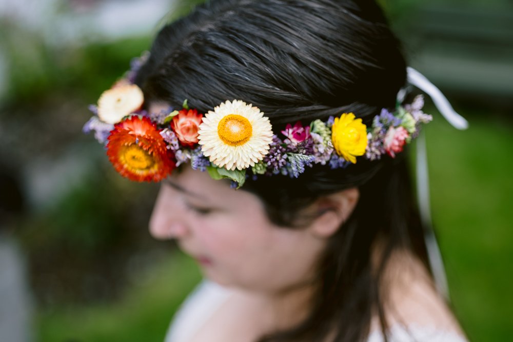 A person with dark hair wears a colorful floral crown featuring orange, yellow, and purple flowers. The background is blurred greenery.