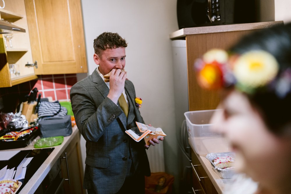 A man in a suit eats a sandwich in a kitchen, holding a napkin. Another person with a flowered accessory is in the foreground.