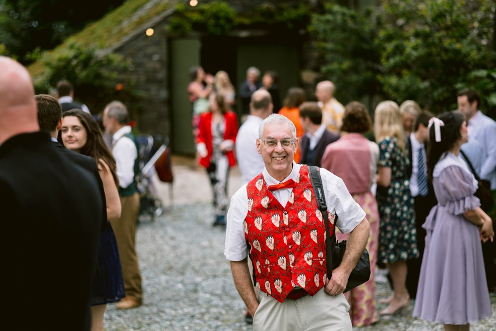 A man wearing a festive red vest with a camera over his shoulder smiles at the camera. A group of people is gathered, some in formal attire, in an outdoor setting.