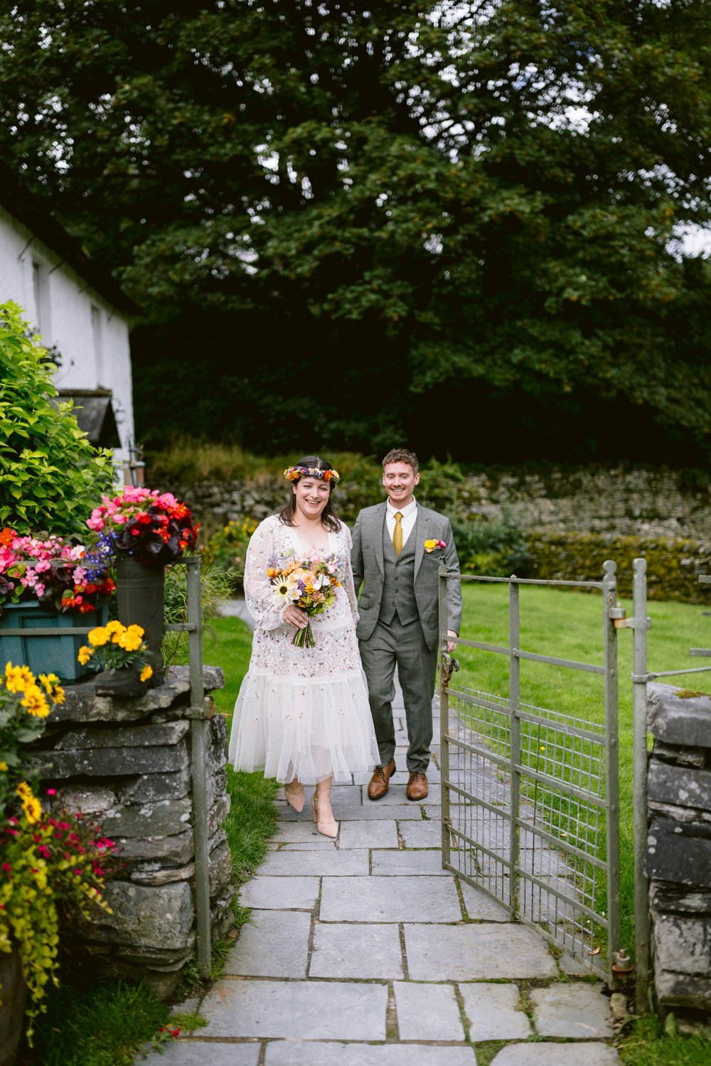 A couple in formal attire walks down a stone path with flowers on each side. The person on the left holds a bouquet and wears a floral headband. They are outdoors near a building and trees.