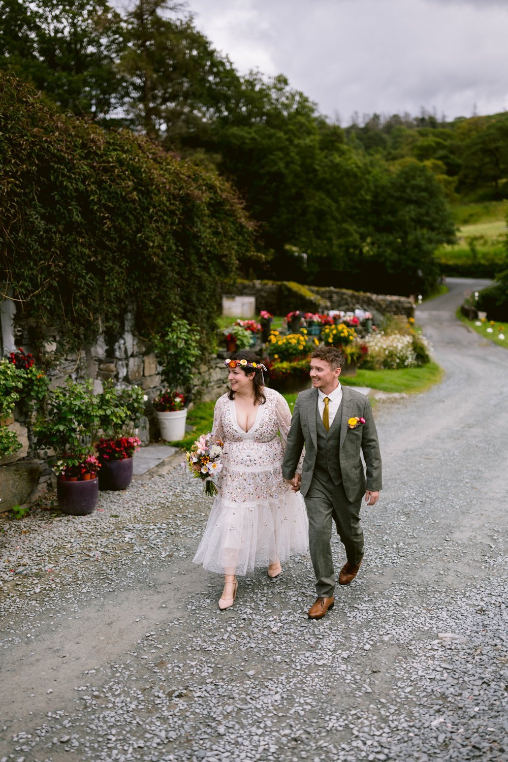 A couple, dressed in wedding attire, walks hand in hand down a gravel path. The woman holds a bouquet. They are surrounded by greenery and colorful potted flowers.