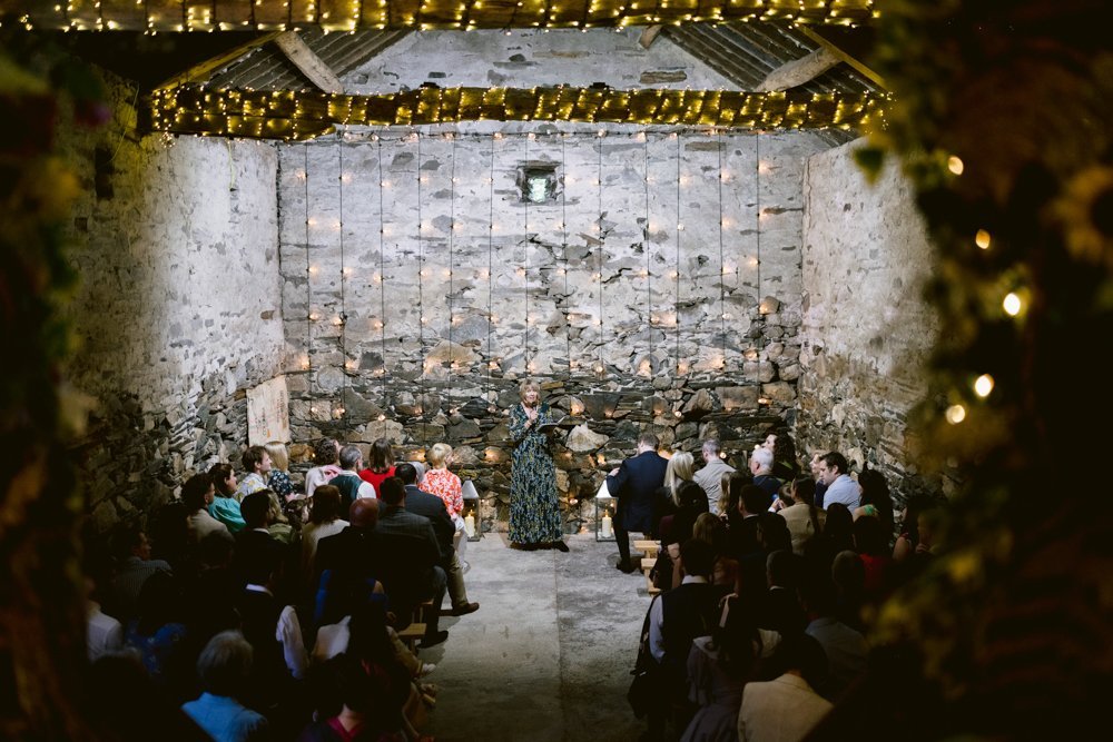 A person stands at the front of a rustic stone room speaking to an audience. The room's ceiling is decorated with string lights.