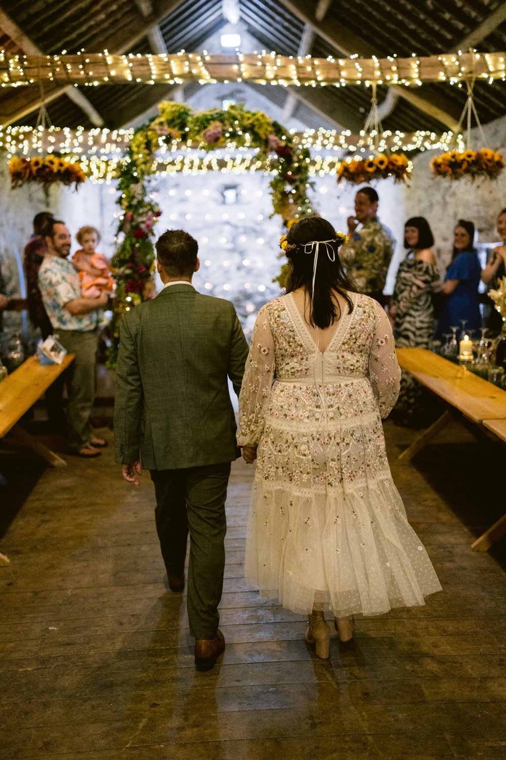 A couple holding hands walks down a floral-decorated aisle in a rustic venue, surrounded by guests.