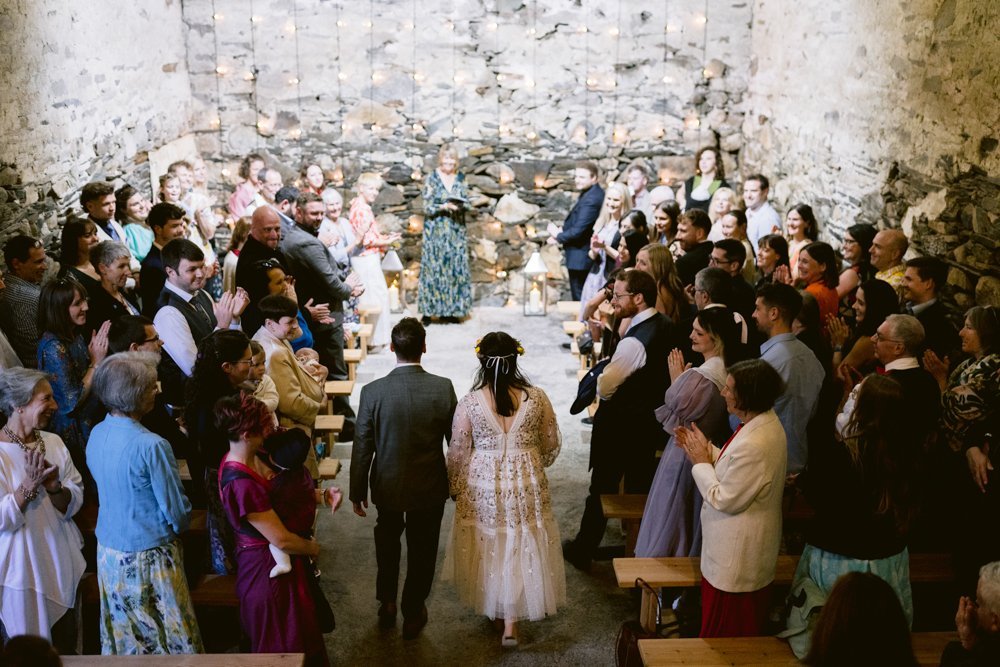 A couple walks down the aisle in a rustic, stone-walled venue. Guests on both sides stand and applaud.