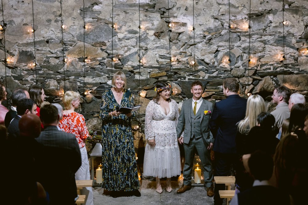 A couple stands holding hands during a wedding ceremony in front of a stone wall with string lights. An officiant reads from a book, while guests are seated on wooden benches.