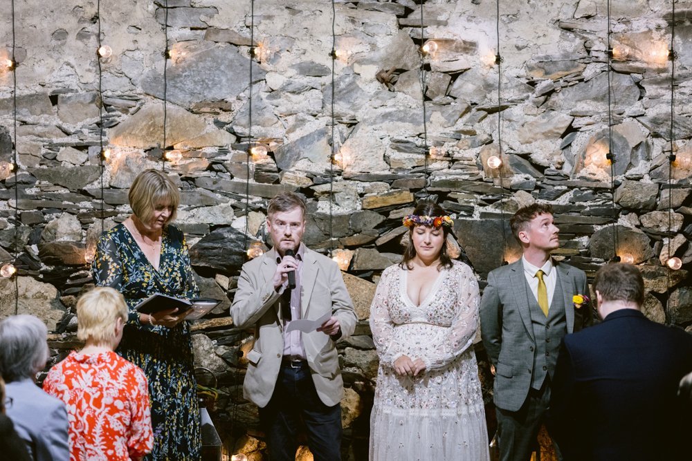 A wedding ceremony with a bride and groom standing by a stone wall. A man is speaking into a microphone, and a woman is holding a book. Guests are seated in the foreground.