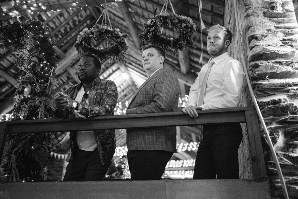Three men standing on a wooden balcony, surrounded by hanging wreaths and festive decorations, in a rustic setting.