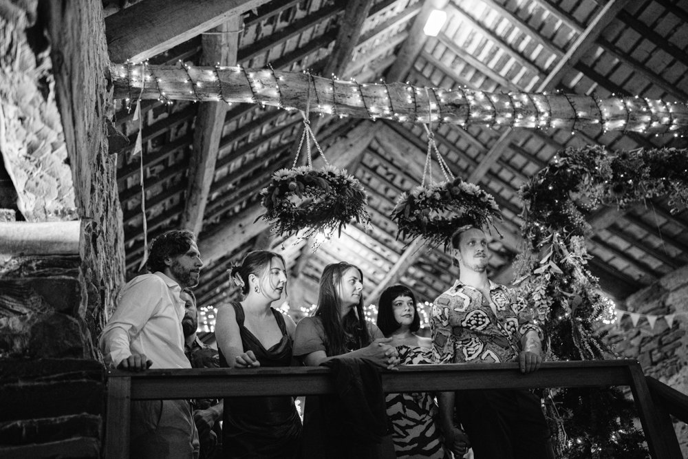A group of people stands on a wooden balcony in a dimly lit room with a high ceiling and hanging lights.