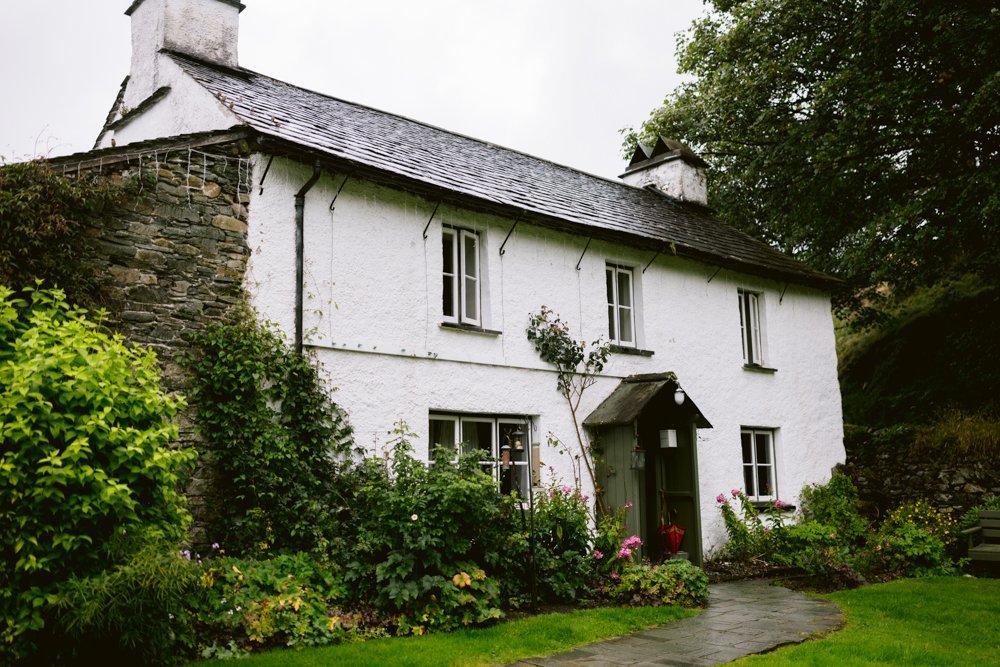 A white stone cottage with a dark slate roof, surrounded by green shrubs and trees. The front yard has a stone path and colorful flowers. Overcast sky in the background.