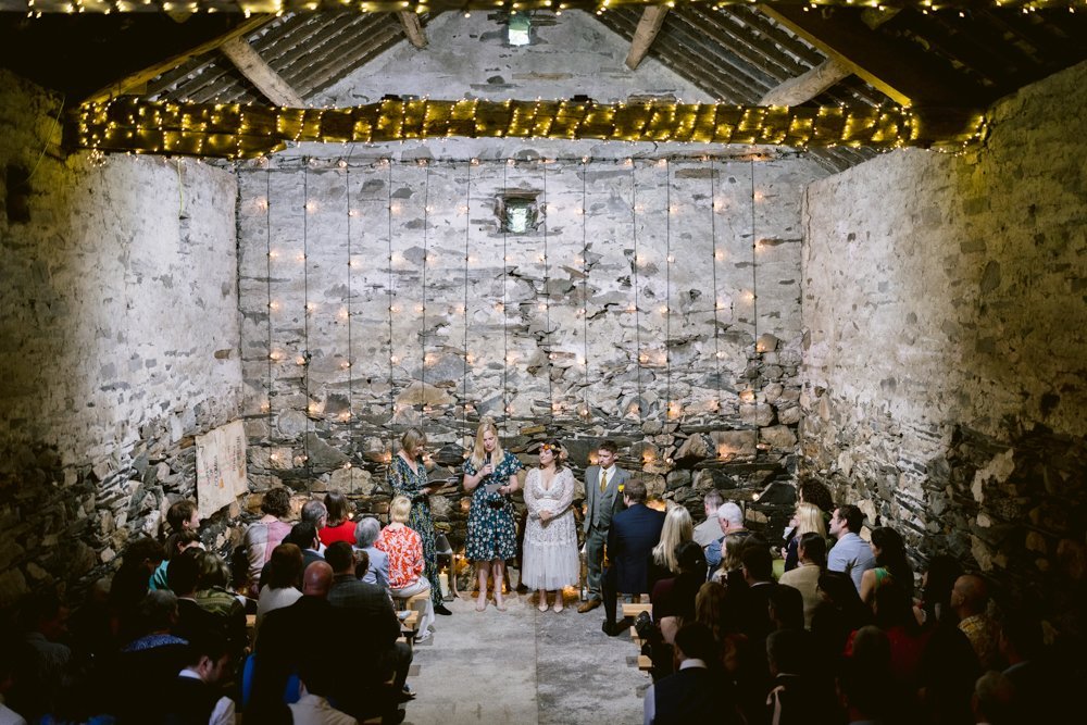 A group of people gathered in a rustic stone barn with string lights on the ceiling, some standing at the front and others seated, as they appear to watch or participate in a ceremony or presentation.