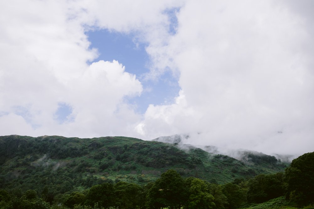Cloudy sky with a small patch of blue over a green, forested hillside.