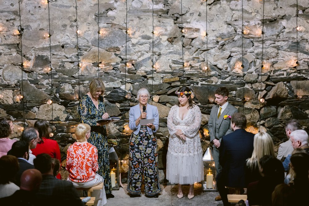 A wedding ceremony in a rustic stone-walled venue with string lights. A couple stands with an officiant and another person, facing seated guests.