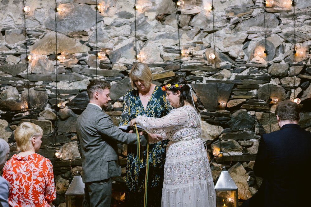 A couple stands facing each other during a wedding ceremony with a stone wall and string lights in the background, as an officiant assists them.
