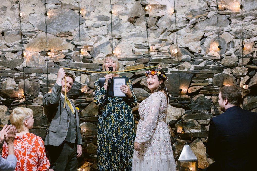 A couple stands in front of a stone wall during a ceremony. The woman officiating holds a scroll. String lights decorate the background, and guests sit nearby.