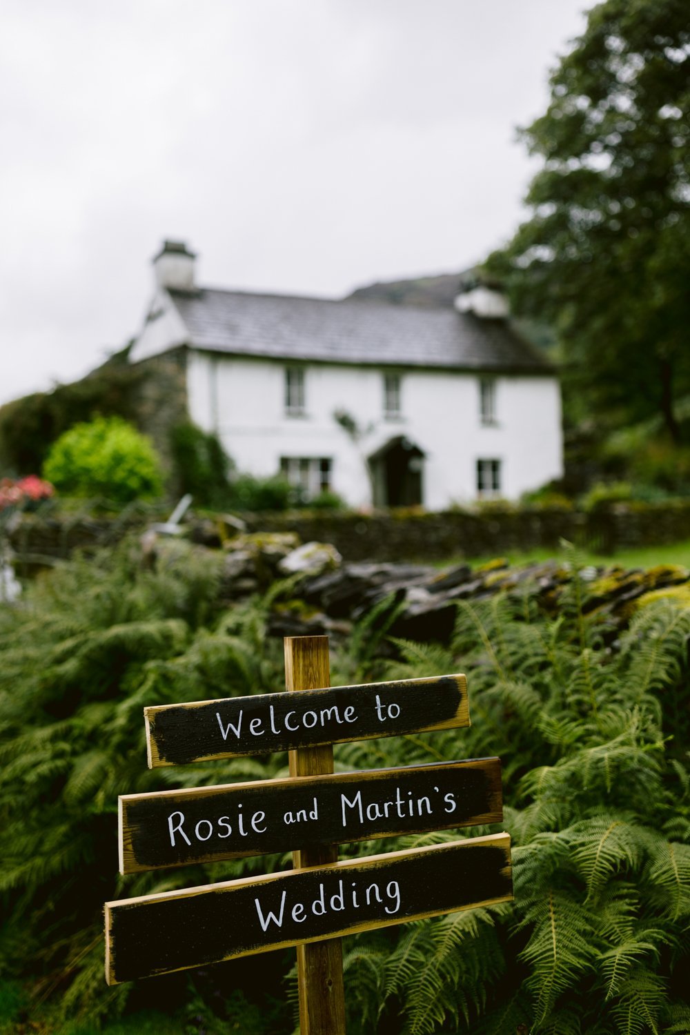 A sign reading "Welcome to Rosie and Martin's Wedding" stands in front of a white house surrounded by greenery.