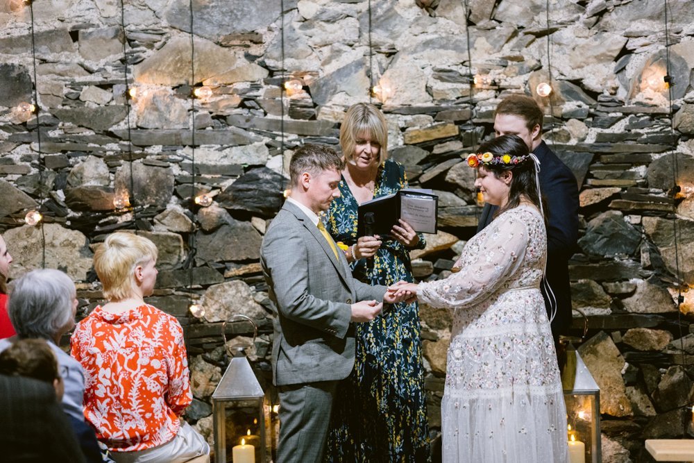 A couple exchanges rings during a wedding ceremony in front of a stone wall adorned with hanging lights. Guests are seated on both sides, and two officiants are present.