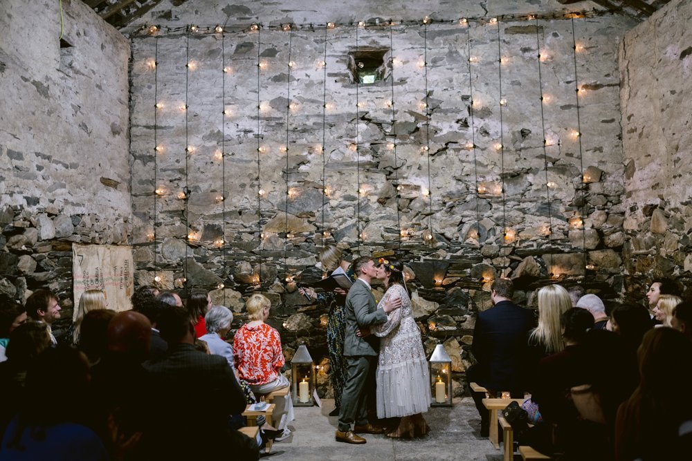 A couple kisses at their wedding ceremony in a rustic stone-walled venue with string lights, surrounded by seated guests.