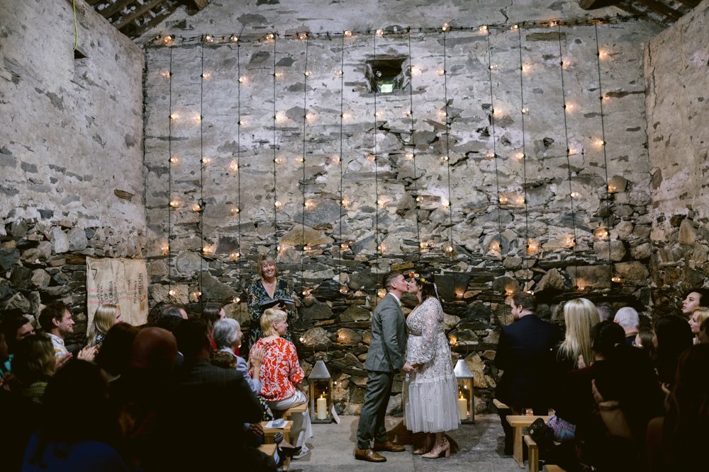 A couple kisses at a rustic wedding ceremony in a stone-walled venue decorated with string lights, surrounded by seated guests.