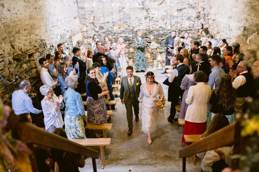 A newlywed couple walks down the aisle, holding flowers, in a rustic venue. Guests seated on wooden benches applaud. Stone walls and string lights are visible.