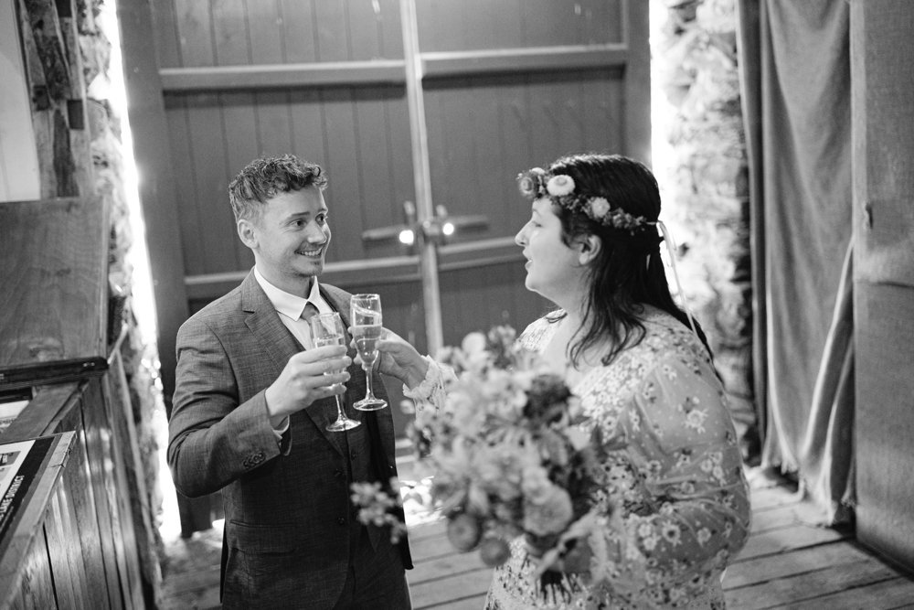 A man in a suit and a woman in a floral dress with a flower crown clink glasses at a celebration, smiling at each other in a dimly lit room. She holds a bouquet of flowers.
