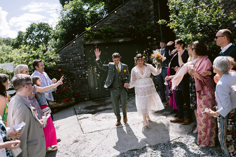 A couple celebrates a wedding, walking through a path where guests are throwing confetti. The bride holds a bouquet, and the groom waves. Both are dressed in formal attire.