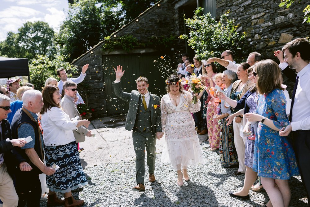 A newlywed couple walks down an outdoor path as guests throw confetti. The bride wears a white dress with floral designs, and the groom is in a green suit. Guests are cheering and taking photos.