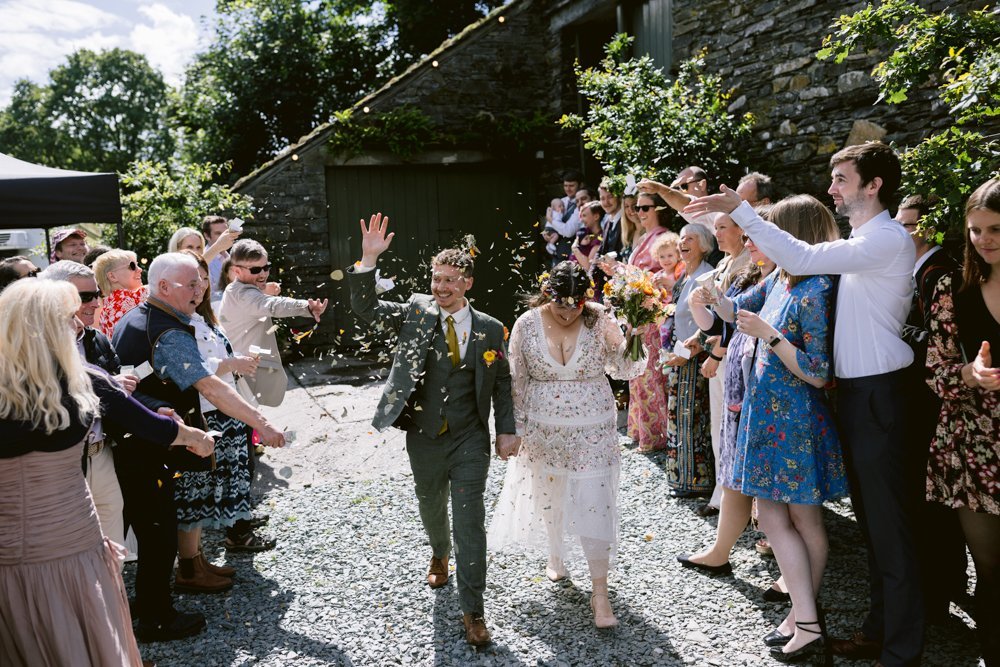 A newlywed couple walks hand in hand down an outdoor aisle as guests throw confetti. They are surrounded by smiling friends and family.