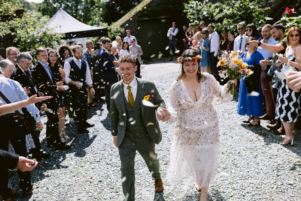 A couple walks happily down a path while guests throw confetti. The woman wears a floral crown and holds flowers; the man is dressed in a suit. People are gathered around, celebrating.