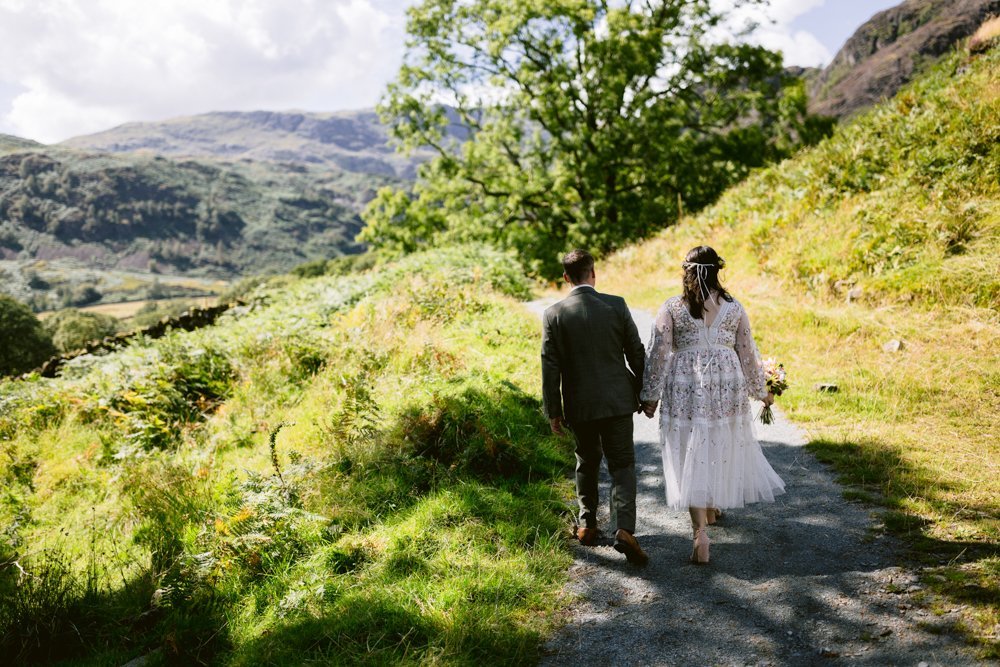 A couple in wedding attire walk hand in hand on a grassy path, surrounded by trees and hills under a partly cloudy sky.
